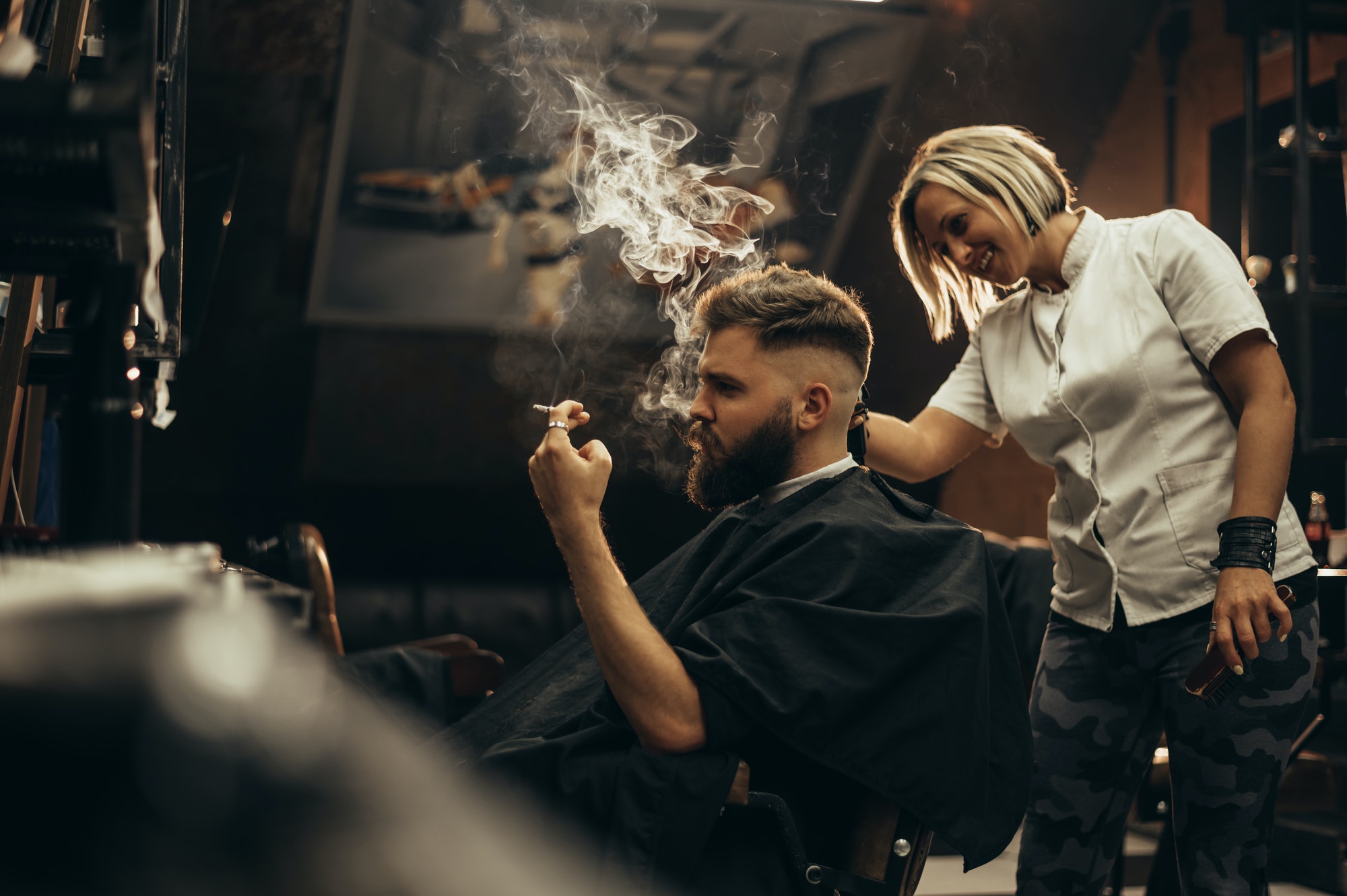 Young bearded man getting haircut by hairdresser and smoking a cigarette